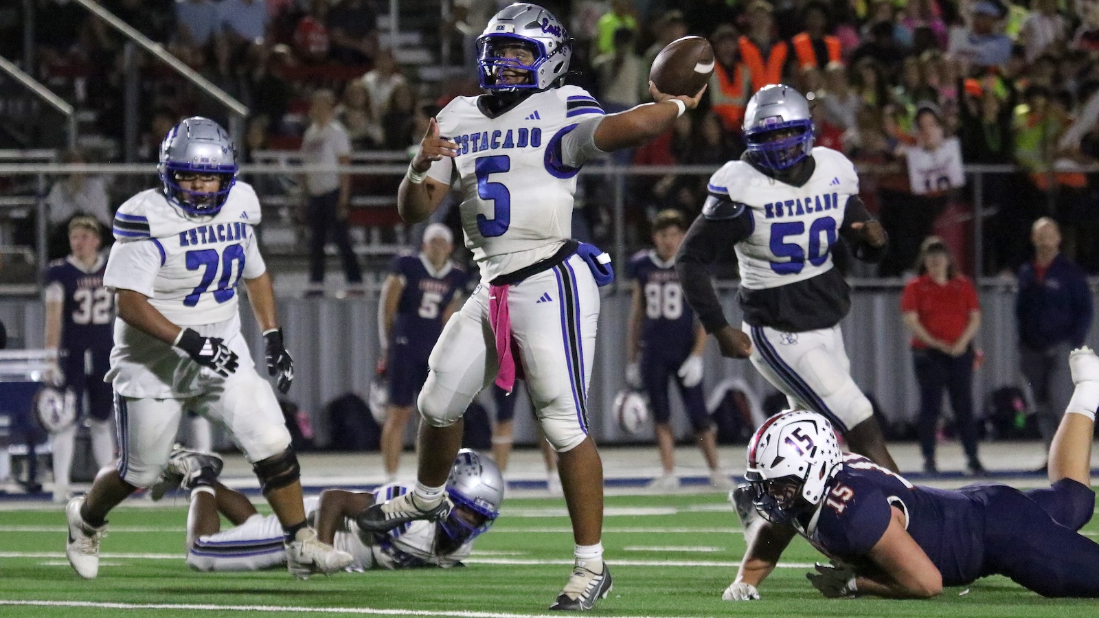 Estacian quarterback #5 in white jersey throws a pass while teammates shield him on the grassy field, crowd cheering in the stands behind.