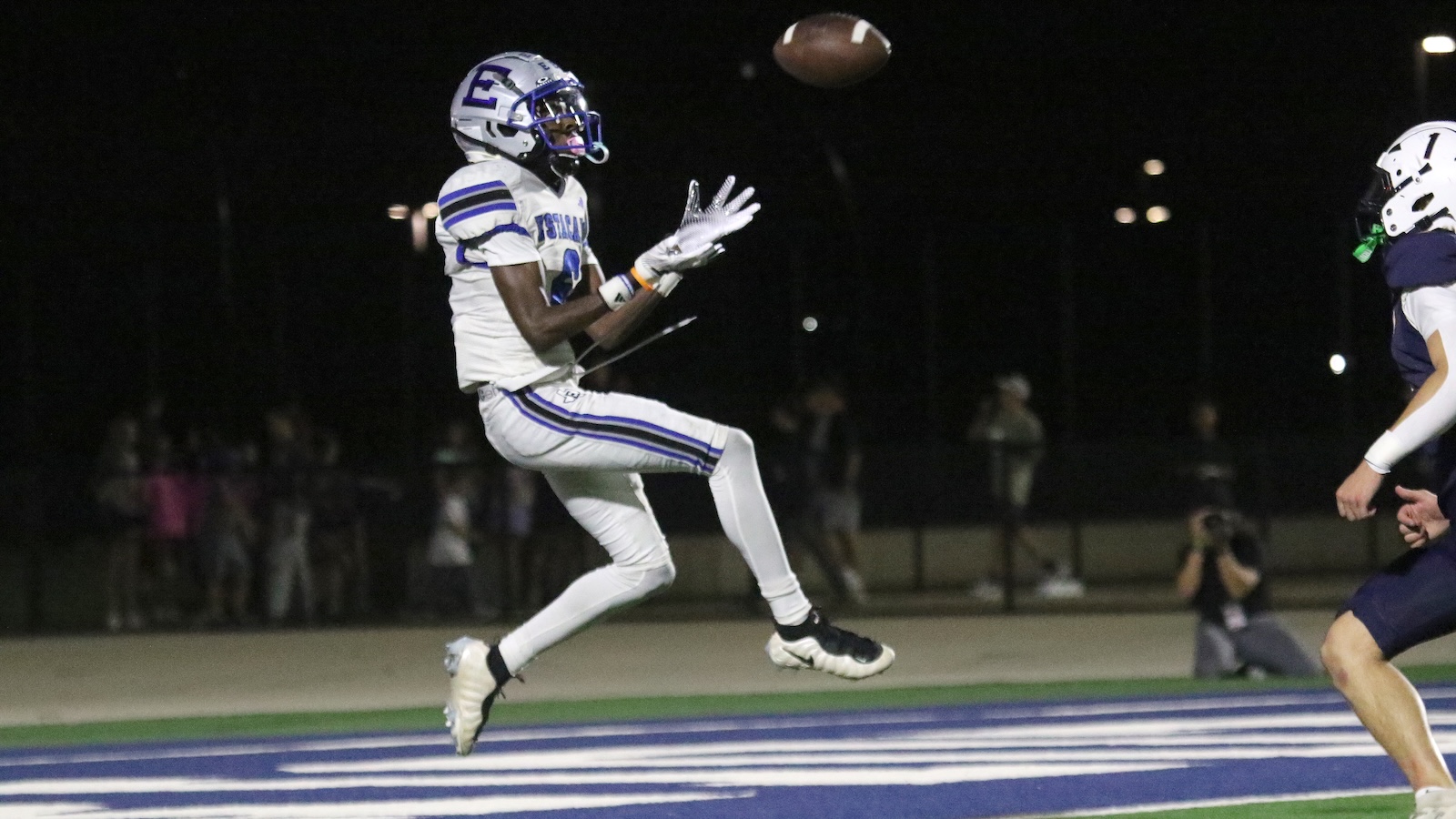 Football player in a white and purple uniform leaps to catch a high pass during a night football game in the end zone area.