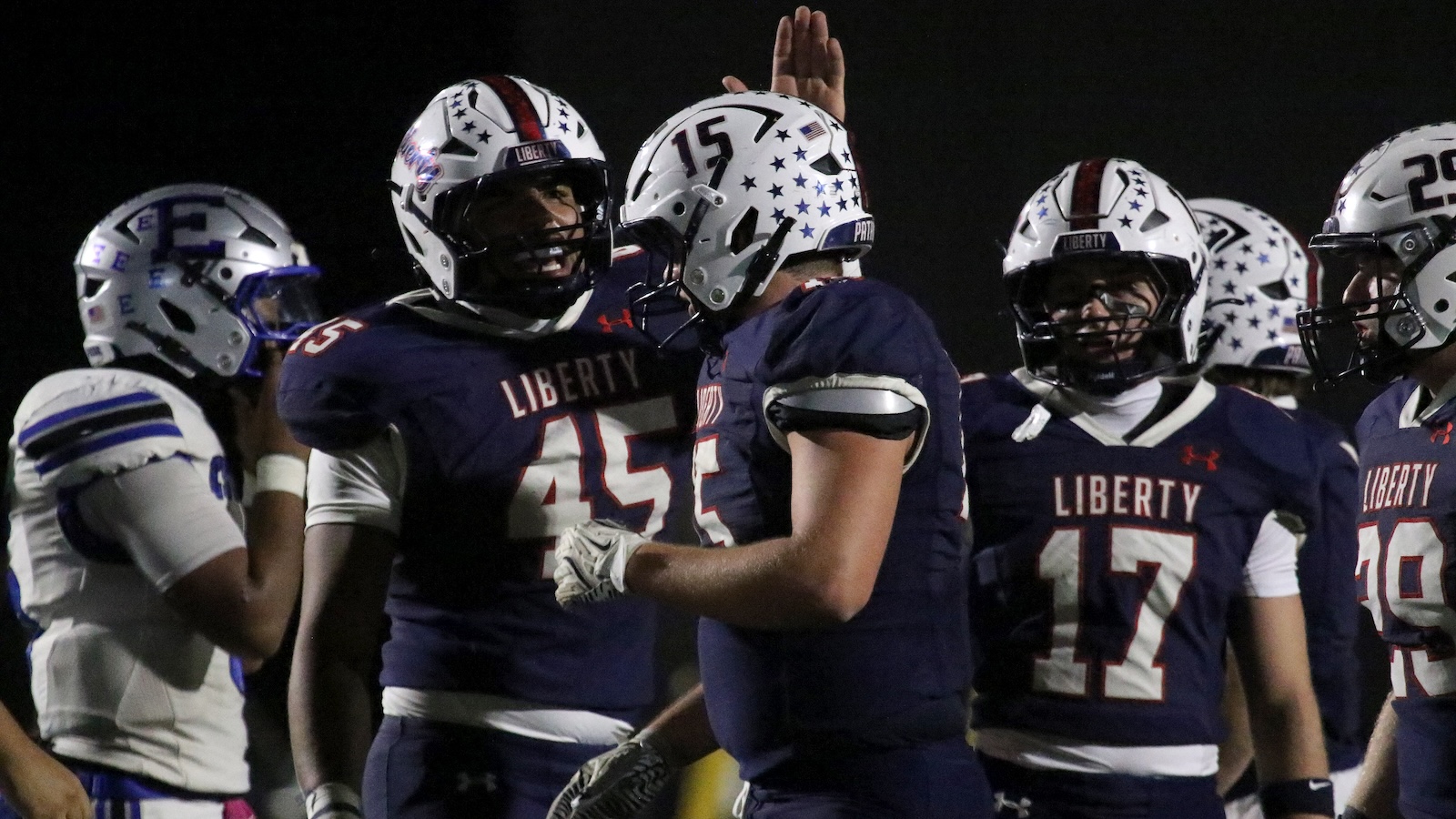 Liberty football players in navy jerseys high-fiving and celebrating on the field at night under stadium lights.