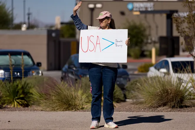 A participant waves while holding a sign reading “USA > Donald Trump” along Eastlake Boulevard during a “No Kings” sidewalk protest organized by Indivisible the 915 in El Paso, Texas, on Saturday, Oct. 17, 2026.
