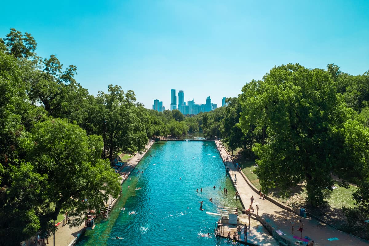 Urban park with a large swimming pool surrounded by trees, city skyline in the background.
