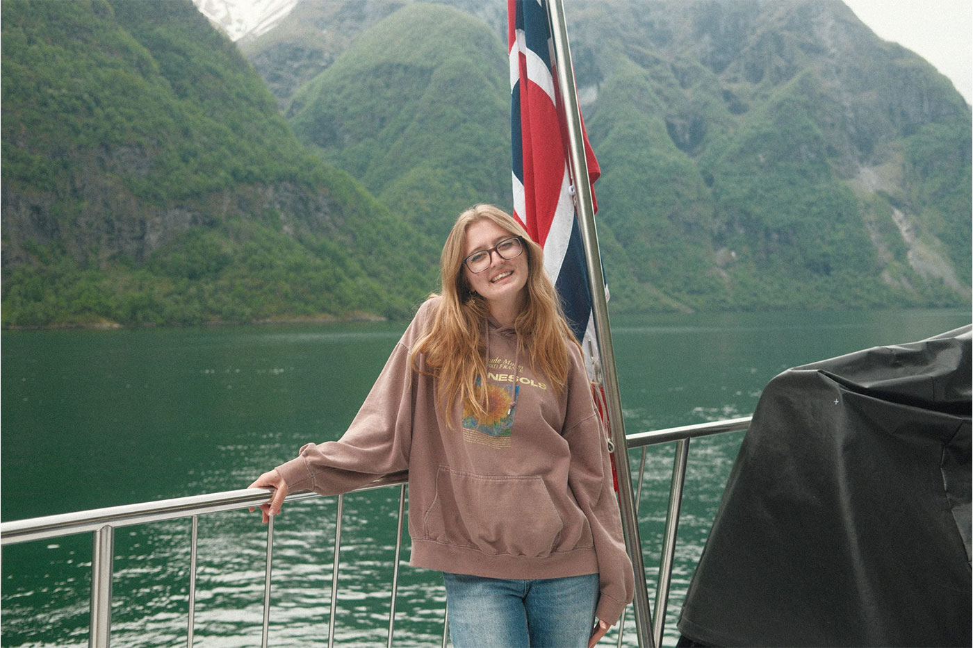 A smiling young woman with glasses stands on a boat in front of a Norwegian flag, with a fjord and steep green mountains in the background.
