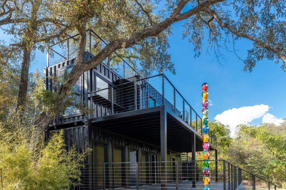 A black, industrial-style building surrounded by trees features balconies and a tall outdoor sculpture composed of stacked, brightly colored faces.
