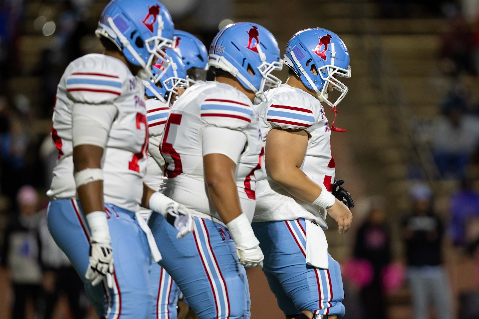 MHS-THS-FB-16 | Press Pass Sports American football players in white jerseys and blue helmets stand in a line on the field, preparing for the play in a stadium.