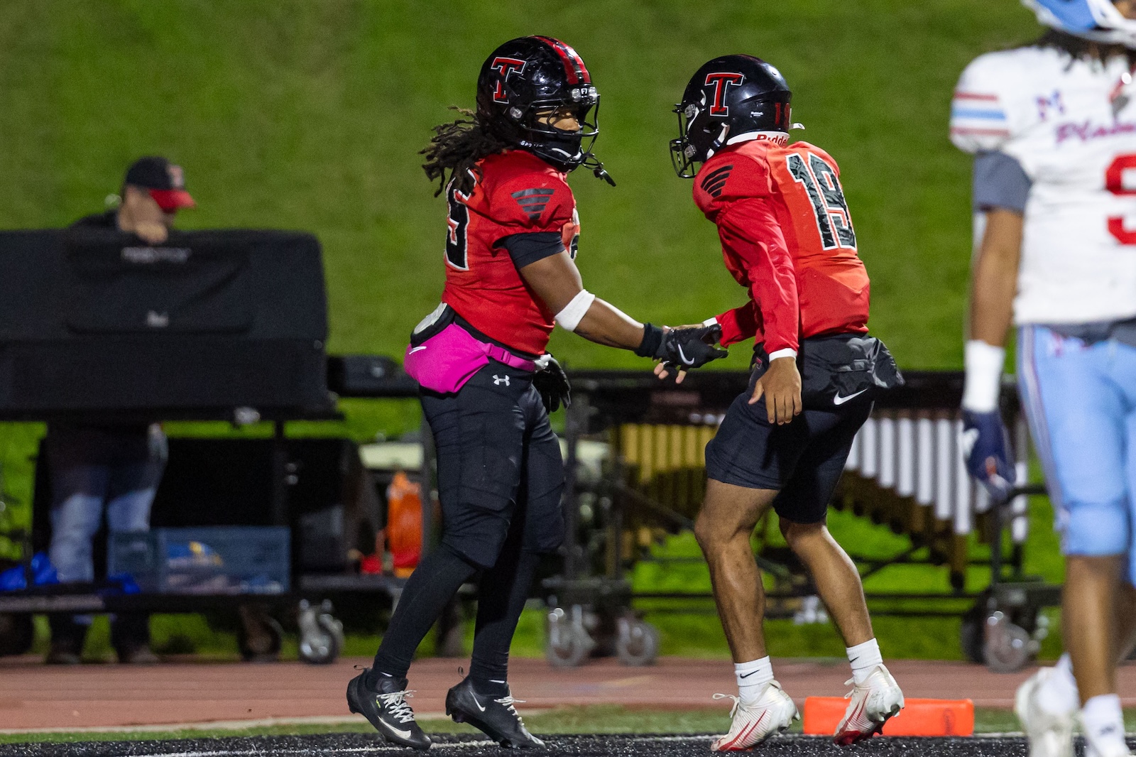 MHS-THS-FB-21 | Press Pass Sports Two football players in red uniforms share a high-five on a track field at night, black helmets with a red T and gloves visible