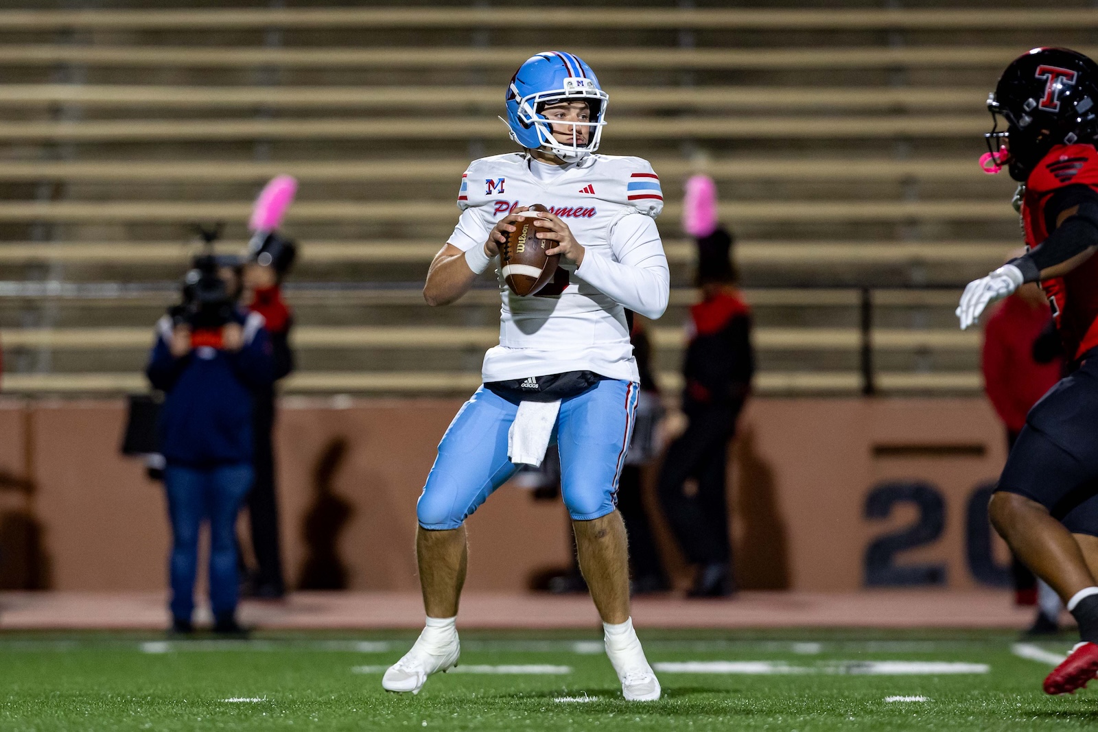 MHS-THS-FB-24 | Press Pass Sports Quarterback in white and blue, gripping the football and looking downfield for a pass on a night game surface.