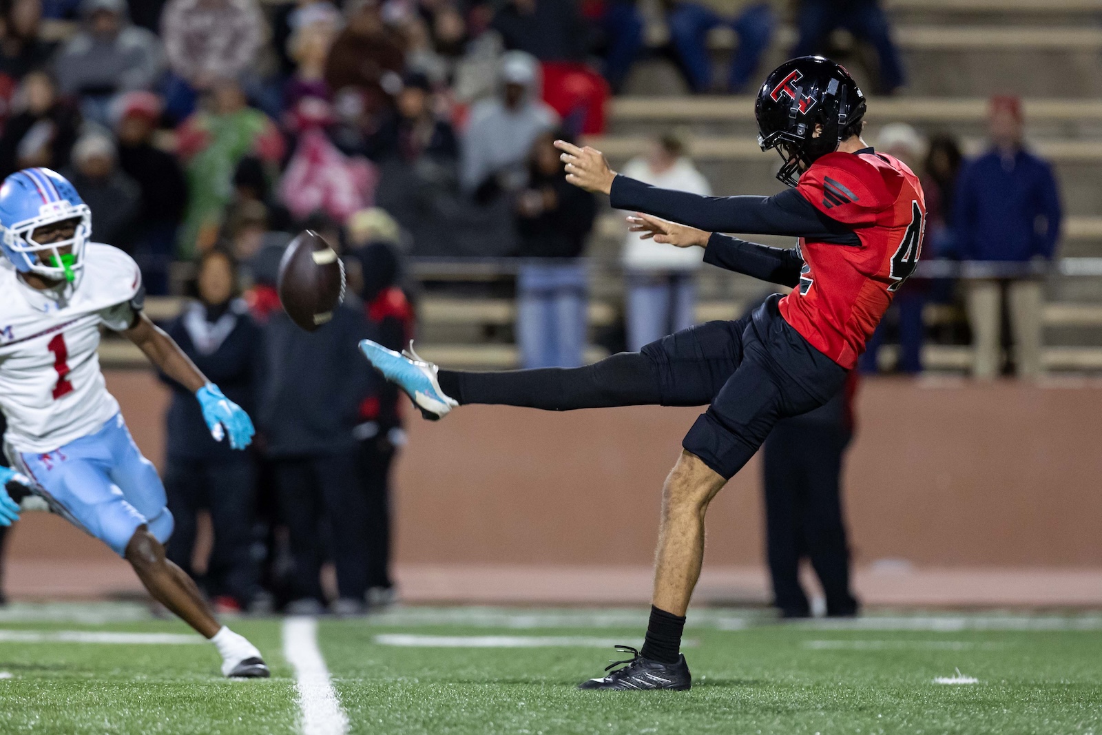 MHS-THS-FB-29 | Press Pass Sports Football game: a player in red jersey kicks a football while an opponent in white and blue pursues from the left; crowd in stands background.