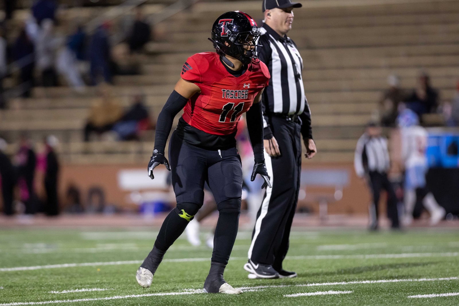 MHS-THS-FB-38 | Press Pass Sports Football player in a red jersey with number 11 and black helmet on a football field, awaiting a play, referee nearby in stripes behind.