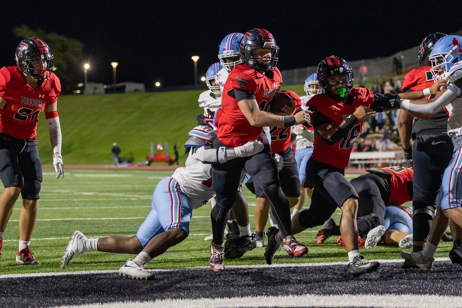 MHS-THS-FB-39 | Press Pass Sports Running back in a red jersey salvages the ball and charges forward as defenders close in during a night football game on a lit field.