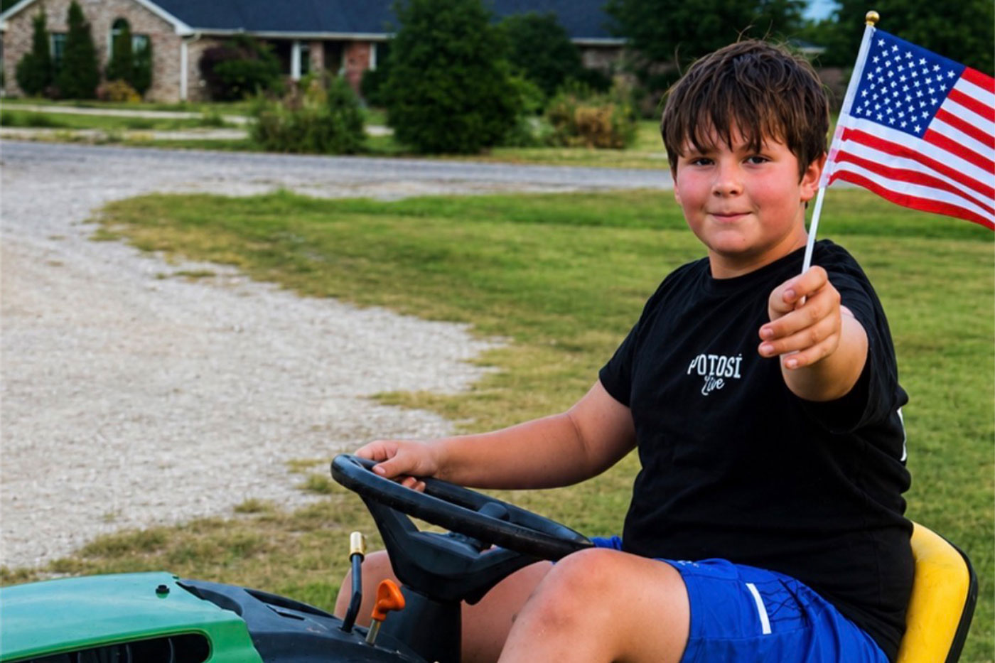 Ty sits atop a green riding mower, holding a small American flag. 