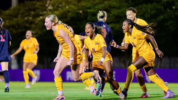 Group of LSU women's soccer players in yellow uniforms cheer and run on the field at night under stadium lights.