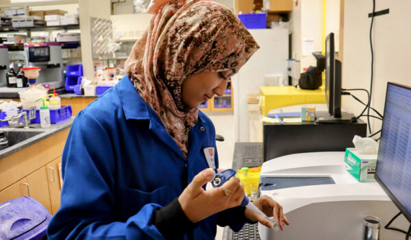 A lab technician in a blue coat examines a small sample at a laboratory bench.