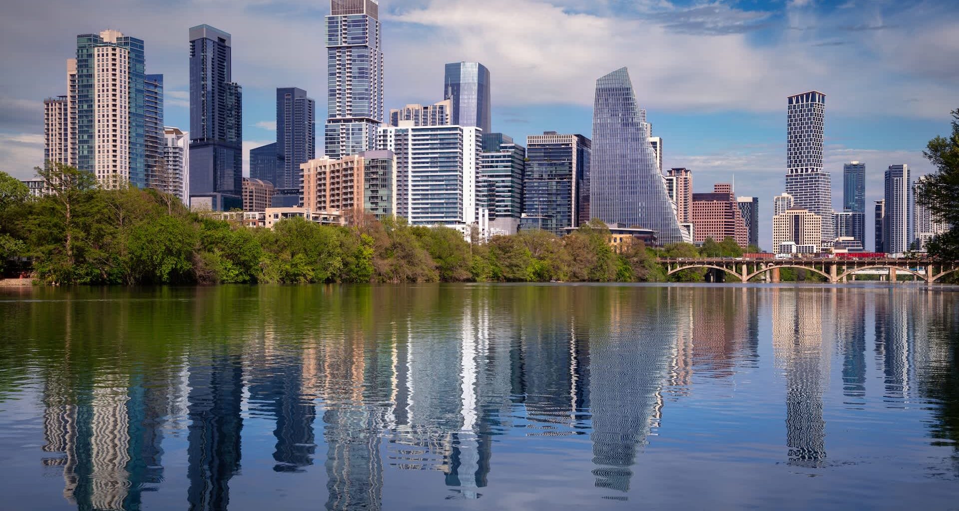 An Austin, Texas, skyline of modern skyscrapers reflected in a calm body of water with greenery in the foreground.