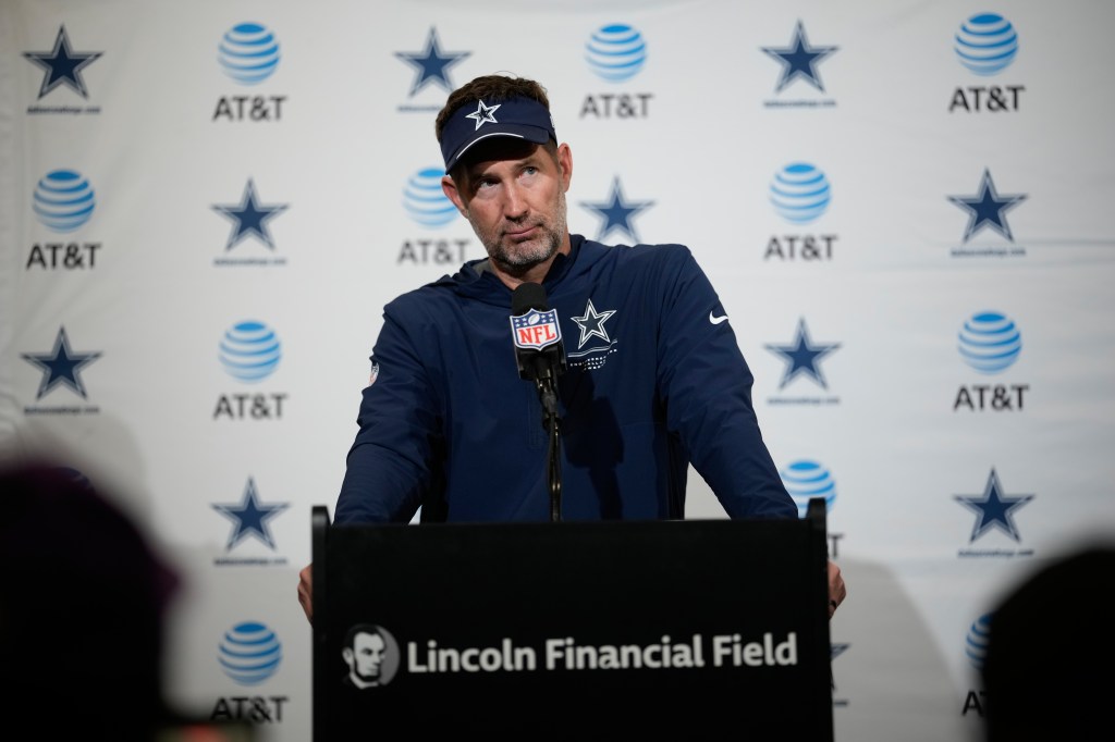 Dallas Cowboys offensive coordinator Brian Schottenheimer speaks at a podium during a news conference in front of a backdrop with AT&T and team logos.