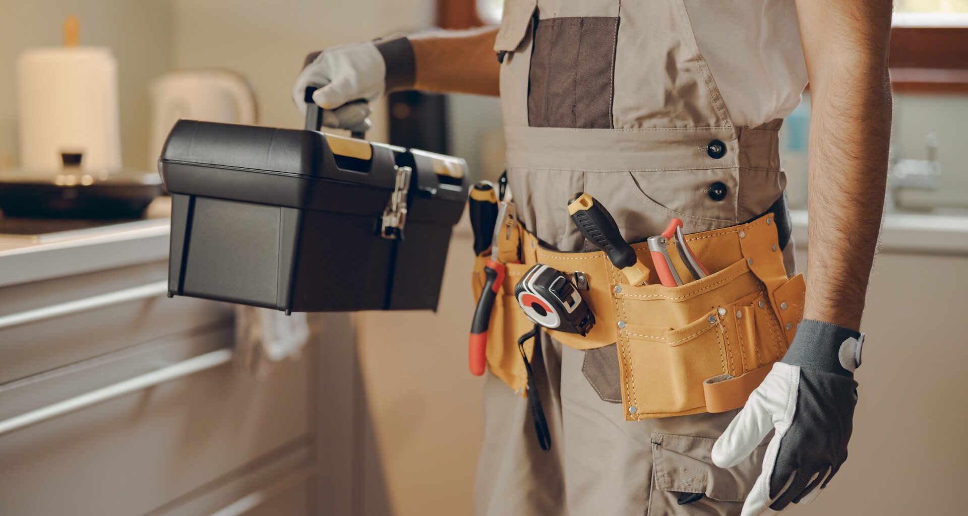 A person holding a toolbox and wearing a tool belt in a kitchen setting.