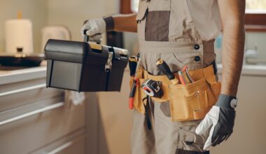A person holding a toolbox and wearing a tool belt in a kitchen setting.