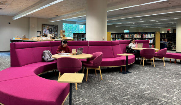 Two students study at round tables within a curved magenta seating area in a library, surrounded by bookshelves, open floor space and large windows providing natural light.