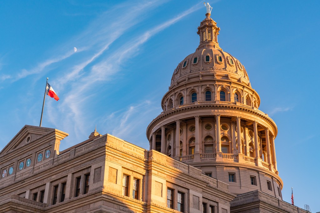 The Texas State Capitol building in Austin, Texas, featuring its prominent dome bathed in golden light against a blue sky with the Texas state flag flying.