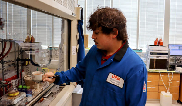 A man in a blue lab coat writes on a glass inside a lab.