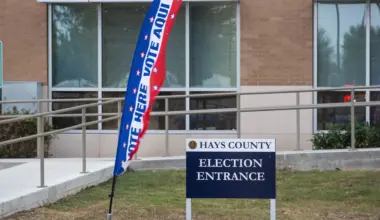 A sign in front of a building reads "Hays County Election Entrance". Beside it, a flag reading "Vote Here" and "Vote Aqui"