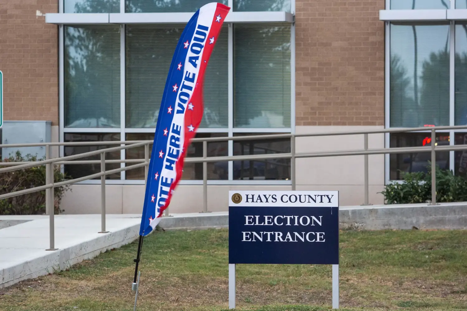 A sign in front of a building reads "Hays County Election Entrance". Beside it, a flag reading "Vote Here" and "Vote Aqui"