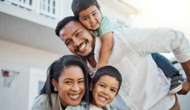 Smiling family of four outdoors, with a man giving a piggyback ride to a young boy, while a woman leans forward beside them, holding another smiling boy in front.