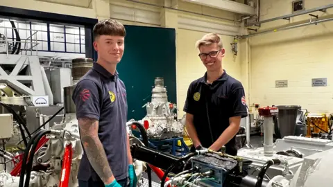 BBC Two men in a factory smile at camera. There are various engine parts behind them. Both are wearing blue tops, and are younger workers. The factory walls are painted yellow.
