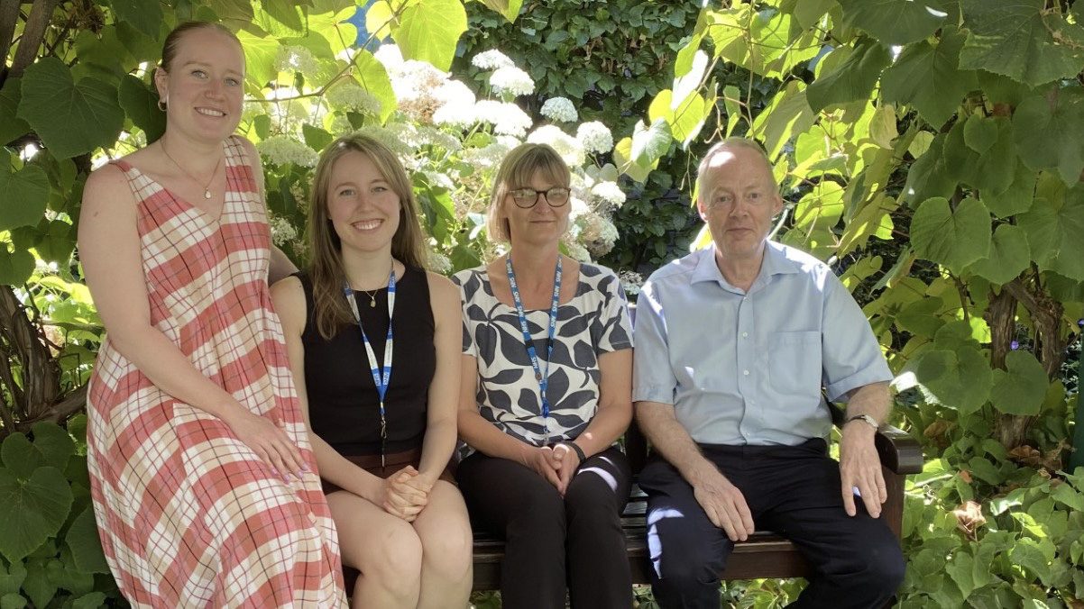 Four people sitting on a bench under a tree, looking at the camera smiling