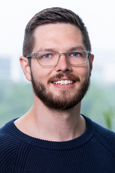 Smiling man with brown hair and facial hair, wearing glases and a dark sweater.