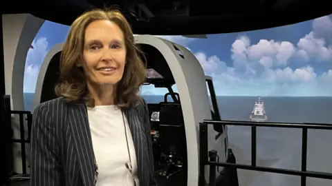 A woman in a pin stripe blue jacket and a white top, smiles at camera. There is a flight simulator behind her.