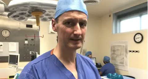 BBC Mr Colbert stands in a hospital operating room, wearing blue scrubs and surgical hat. Two members of staff, also in scrubs, can be seen over his left shoulder looking at a screen.