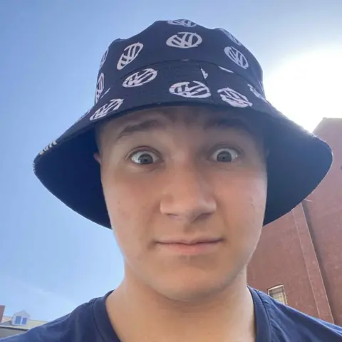 Oliver Holton A selfie of a man in a black and white patterned bucket hat. He is stood outside and in the background there is a building and the sky