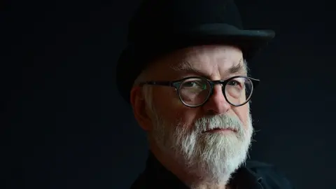 Getty Images Portrait of Terry Pratchett, who is wearing a black shirt and black brimmed hat, in front of a plain black background. He has a white/grey beard and is wearing black glasses.