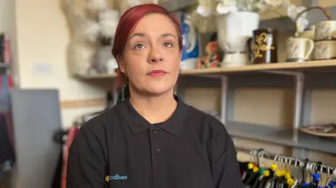 Kerry Ritson is wearing a black polo shirt with the GoodLives logo on. She has her hair dyed dark red. She's sitting in the charity shop which she works.