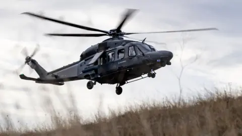 Leonardo Helicopters A helicopter flying. It is black. The camera is looking up. Grass on the floor is blowing from the rotors.