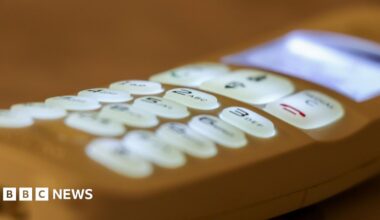 A white man with brown hair standing outside a house in the rural countryside smiling.