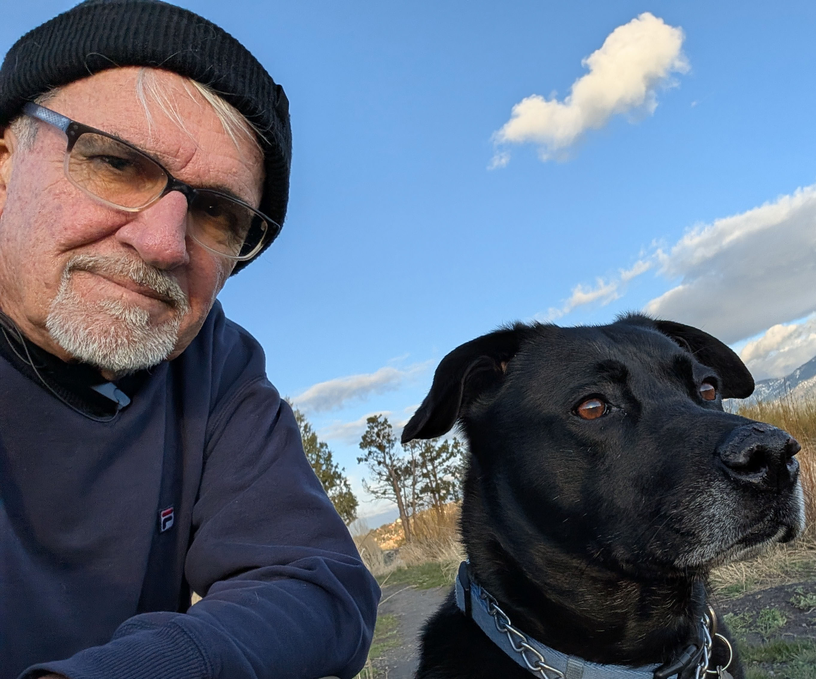 A man wearing a black hat and glasses takes a selfie outside with his black-haired dog