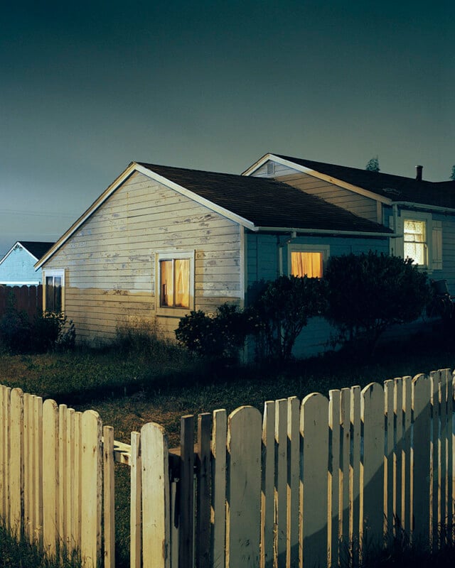 A white wooden house at night with lights glowing from its windows, surrounded by bushes and grass, and enclosed by a white picket fence. The scene is quiet and illuminated by soft, ambient lighting.