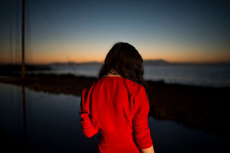 A person with long dark hair, wearing a bright red dress, stands with their back to the camera near a body of water at sunset, with the sky fading from orange to deep blue.
