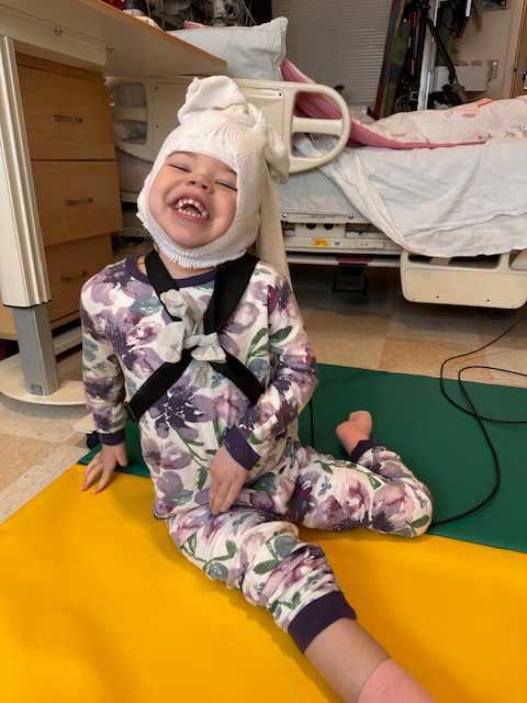 In a hospital room, Juniper Adkisson, age 4, smiles for a photo while hooked up to an electroencephalogram to monitor her brain waves for seizure activity. Despite trialing over a dozen medications and undergoing multiple surgeries, Juniper still experiences daily seizures due to an undiagnosed rare disease.