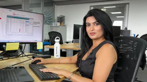 A woman in a black top, with black hair, sits at a desk. She is looking into the camera. Her hands are on a keyboard and mouse, and a computer monitor is behind her.