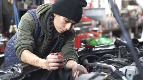 Getty Images A woman with long black hair in two plaits wearing a black beanie hat, khaki hoodie and navy quilted bodywarmer working as a mechanic with her oil covered hands inside the engine of a car with its bonnet up