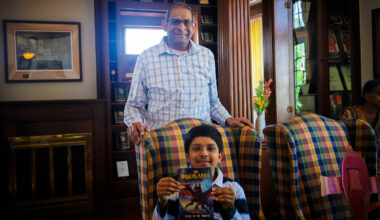 A proud father stands behind his 11-year old son. The son holds his original novel, a paperback book called "Secret of the Dragons."