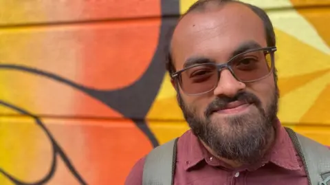 BBC / Elaine Dunkley A young Asian man with brown hair and a beard smiles down the camera, to the right of frame. He is wearing sunglasses, a purple shirt and a backpack. He is stood in front of a colourful wall with bright orange graffiti on it. 
