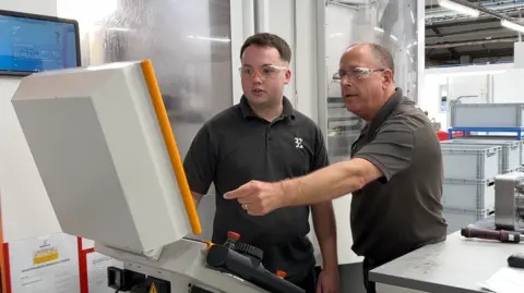 Two men in t shirts look at a screen in a factory. One is older. The older man is pointing at the screen.
