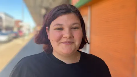 BBC / Kate McGough Libby smiles into the camera in front of a row of shuttered shops on a sunny day in Grimsby. She has cropped red hair and is wearing a black top.