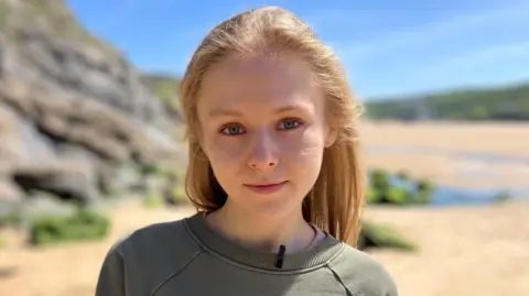 BBC / Dan Nelson A blonde young woman with blue eyes and wearing a grey sweatshirt looks into the camera. She is standing on a beach in Cornwall and there is blue sky behind her. 