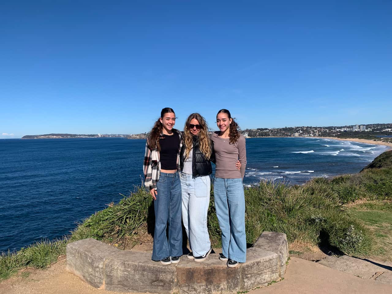 A woman stands between two teenage girls on a cliff, with the ocean behind them.