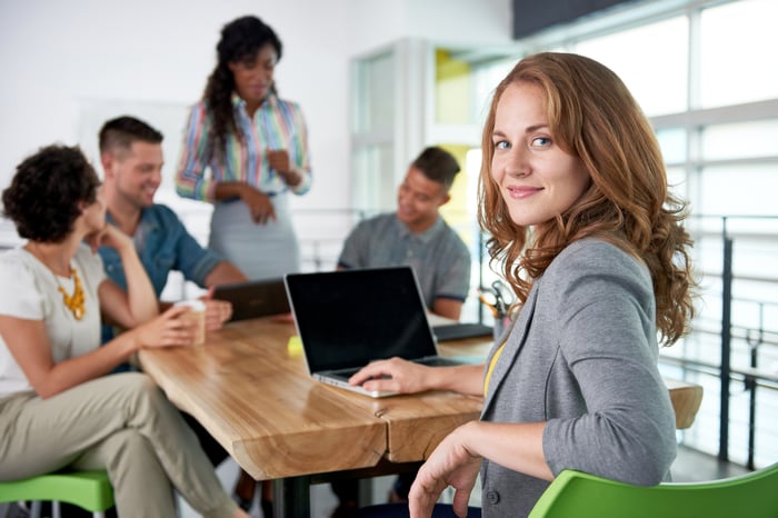 At a table with several laptops and people talking, one person looks back and smiles at the camera.