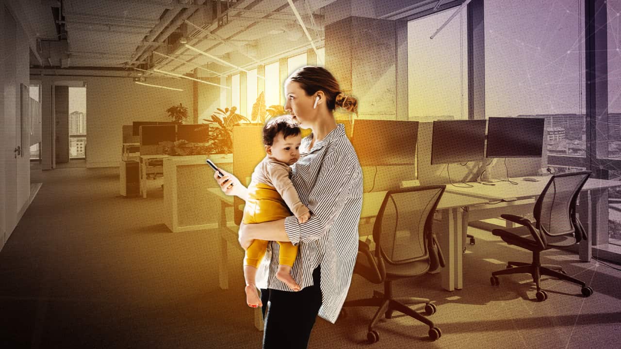 A woman holds a baby while looking at her mobile phone, against a background of an office environment.